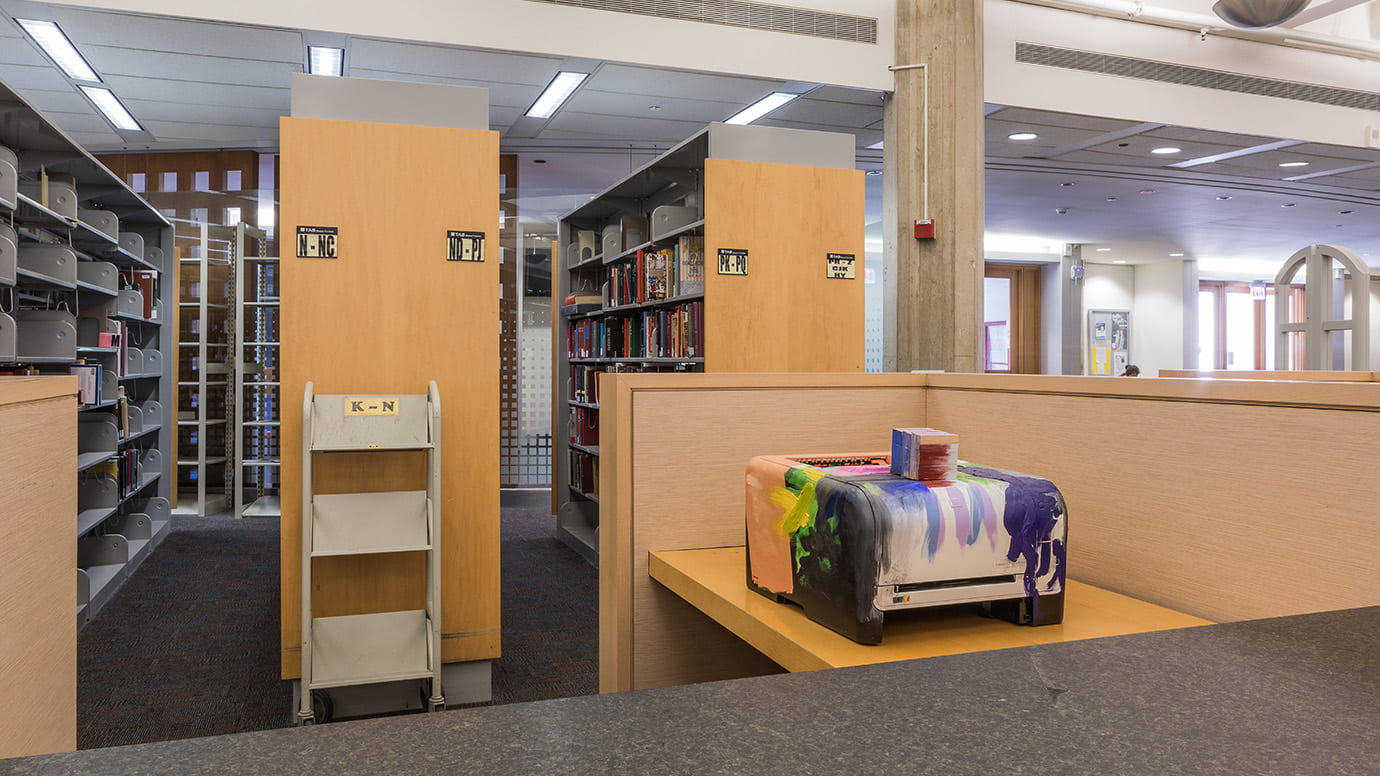 A printer that has been made into a sculpture sits in a cubicle near a stack of books